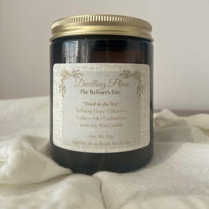 May include: A brown glass jar candle with a gold lid. The label reads "Dwelling Place" and "The Refiner's Fire." The label also lists the scent notes: Cedar, Ash, and Cashmeran. The candle is 100% soy wax and has a burn time of approximately 18-25 hours.