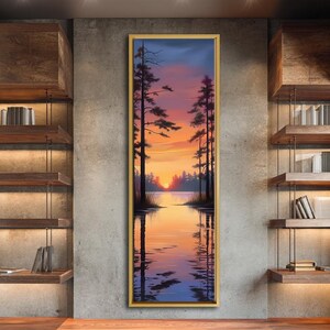Op de afbeelding: Een ingelijst landschapsschilderij toont een zonsondergang boven een meer, met silhouetten van bomen. De lucht verandert van blauw naar oranje en rood, weerspiegeld in het water. Het kunstwerk hangt aan een betonnen muur.