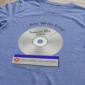 May include: A pale blue t-shirt featuring a graphic with the text "Disc Write Error" and "Summer Mix 2002." The design includes a cracked CD and an error message. A computer mouse and CD cases are also visible.