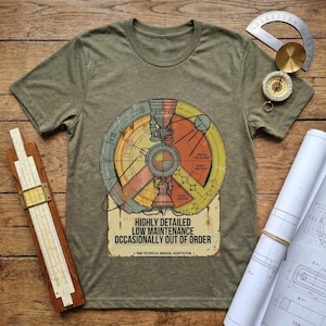 May include: Olive green t-shirt featuring a colorful diagram with text that reads "HIGHLY DETAILED LOW MAINTENANCE OCCASIONALLY OUT OF ORDER." The diagram has orange, yellow, blue, and green sections. A slide rule, compass, and blueprints are on a wooden surface.
