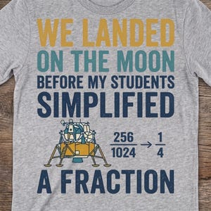May include: A heather grey t-shirt featuring the text WE LANDED ON THE MOON BEFORE MY STUDENTS SIMPLIFIED A FRACTION in yellow, teal, and dark blue. Below the text is a graphic of a lunar lander next to the math equation 256/1024 equals 1/4.