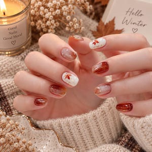 May include: Manicured fingernails featuring a seasonal autumn theme with a mix of glossy solid deep reddish-brown polish, translucent ombre gradients, and white accent nails decorated with small, detailed red and orange maple leaf decals.