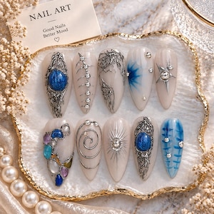 May include: Ten almond-shaped press-on nails featuring silver metallic accents, blue stones, and star motifs. A card labeled NAIL ART Good Nails Better Mood is visible. The designs include textured silver filigree, metallic spheres, and blue painted patterns on a neutral base.