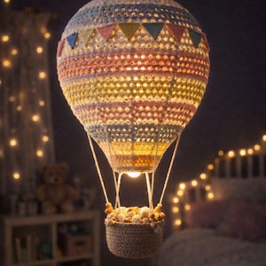 Op de afbeelding: Een handgemaakte gehaakte luchtballamp met een kleurrijk gestreept ontwerp. De lamp heeft een mandje gevuld met decoratieve elementen en hangt aan het plafond. Het warme licht creëert een gezellige sfeer.