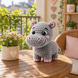 Pode incluir: Um hipopótamo de crochê cinza com detalhes rosa nas orelhas, bochechas e pés. O brinquedo de pelúcia feito à mão tem olhos grandes e pretos e uma expressão sorridente. Está em cima de uma mesa de madeira.