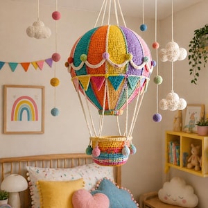 Könnte beinhalten: Eine farbenfrohe, gehäkelte Heißluftballon-Lampe hängt von der Decke und weist leuchtende Streifen in Orange, Gelb, Grün und Lila auf. Die Lampe hat einen Korb und wird von weißen Schnüren gehalten. Dekorative Pompons und wolkenförmige Ornamente ergänzen die Kinderzimmerdekoration.