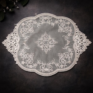 May include: An off-white, oval lace doily featuring detailed floral and scroll patterns. The delicate lace has a scalloped edge and a central design of swirling motifs. Ideal for home decor, approx. 30 cm in length.