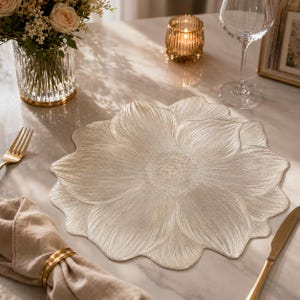 Puede incluir: Un mantel individual beige en forma de flor con un diseño detallado de pétalos. El mantel individual está sobre una superficie de mármol, con un tenedor, un cuchillo y un servilletero dorados. También hay una copa de vino, una vela y un jarrón con flores.