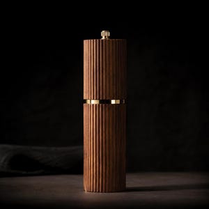 May include: A tall, wooden pepper mill with a ribbed design. The dark brown wood is accented with a gold band and a gold top. The mill is cylindrical and stands upright against a dark background.
