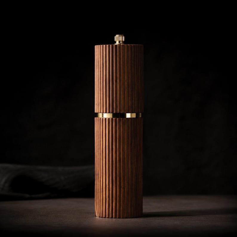 May include: A tall, wooden pepper mill with a ribbed design. The dark brown wood is accented with a gold band and a gold top. The mill is cylindrical and stands upright against a dark background.