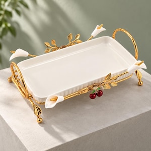 May include: A rectangular white ceramic tray set within a gold-toned metal frame. The frame features decorative gold leaves, white calla lily accents, and a small pair of red cherries. The tray is supported by two arched gold handles and feet.