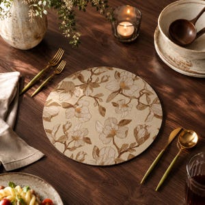 May include: A round beige placemat with a floral design featuring brown magnolia flowers and leaves. The placemat is set on a dark wooden table, alongside gold-colored cutlery, a candle, and a bowl.