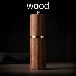 May include: A wooden pepper mill with a ribbed design. The mill is a dark brown color with gold-colored accents. The word "wood" is written in white at the top of the image. The mill is a kitchen utensil.