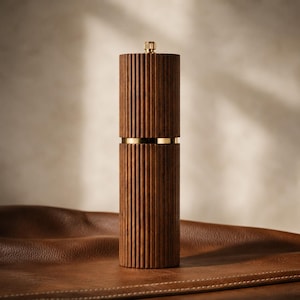 May include: A wooden pepper mill with vertical grooves and gold-colored accents. The top and base are cylindrical, with a gold band separating the two sections. The mill has a gold-colored top and sits on a brown leather surface.