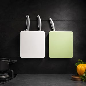May include: Two square wall-mounted knife blocks, one white and one light green, each featuring a single silver metal knife handle protruding from the top. The blocks are mounted side-by-side on a dark, textured wall surface in a kitchen setting.
