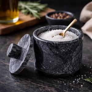 Puede incluir: Un salero de mármol gris oscuro con tapa a juego y una pequeña cuchara dorada. El recipiente está lleno de sal gruesa blanca. La tapa está ligeramente entreabierta, revelando el contenido. El fondo incluye una tabla de cortar de madera y un cuenco pequeño.