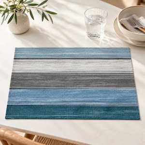 May include: A rectangular placemat featuring horizontal stripes in varying shades of blue, gray, and white. The design has a textured, woven appearance with distinct bands of dark teal, medium blue, light gray, and white across the surface.