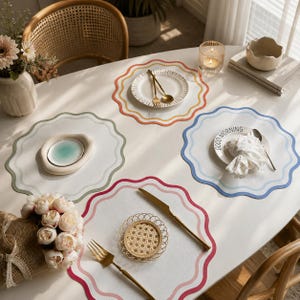 May include: A white oval table set with scalloped placemats in various colors: green, orange, red, and blue. Each placemat has a matching border. Gold-colored cutlery and small plates are arranged on the placemats, creating a dining setting.
