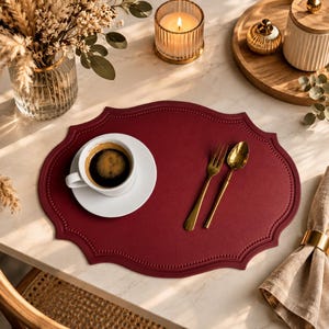 May include: A burgundy placemat with a decorative edge, holding a white teacup and saucer with coffee, a gold fork and spoon. A lit candle, vase of flowers, and napkin with a gold ring are also on the table.