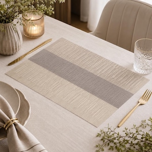 Peut inclure: Un set de table rectangulaire avec une surface texturée et une bande grise centrale. Le set de table est beige et gris, posé sur une nappe claire. Une fourchette et un couteau dorés sont visibles.