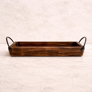 May include: A rectangular wooden tray with a dark brown finish. The tray has two curved, black metal handles on either end. The tray is designed for serving or display purposes.