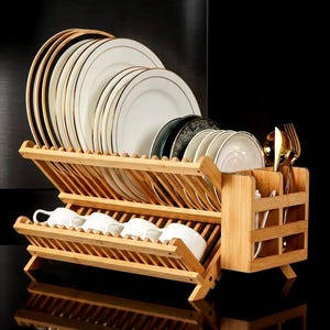May include: A bamboo dish drying rack with multiple compartments. The rack holds white plates with gold rims, cups, and silverware. The rack is made of light brown bamboo and has a slatted design.