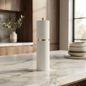 May include: A cylindrical salt or pepper mill featuring a white fluted exterior with vertical ridges. The mill is divided into two sections by a thin, polished gold-colored metal band, with a small gold-colored knob centered on the top cap.