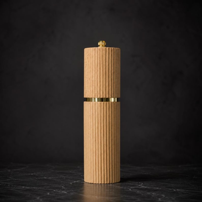 May include: A tall, cylindrical pepper mill with a light brown, ribbed body. The mill features a gold-colored band and a gold-colored top. The design is modern and minimalist, suitable for kitchen decor.
