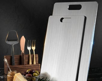 May include: Two stainless steel cutting boards with handle cutouts stand upright. A cheese slicer, cheese knife, and two forks are displayed in a wooden holder. A small bowl of olives, a wedge of cheese, and a jar of mustard are also present.