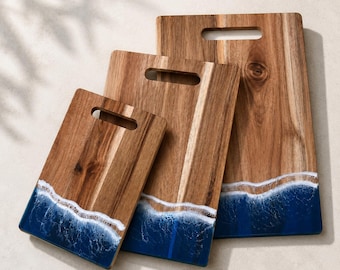 May include: Three rectangular wooden cutting boards of varying sizes, each featuring a natural wood grain finish and a handle cutout at the top. The bottom portion of each board is decorated with a deep blue resin inlay mimicking ocean waves.