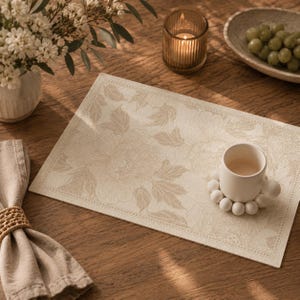May include: A beige rectangular placemat with a floral pattern, a white ceramic cup, and a napkin with a woven ring. A lit candle in a glass holder and a bowl of grapes are also on the wooden table. The placemat has a detailed floral design.