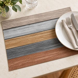 May include: A rectangular placemat featuring horizontal stripes in shades of beige, light gray, tan, dark gray, and brown with a textured fabric appearance. A white plate, a folded beige napkin, and a silver fork and knife rest on the right side.