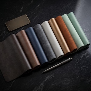Könnte beinhalten: Eine Sammlung von Lederproben in verschiedenen Farben, darunter Braun, Blau, Grau und Grün, diagonal angeordnet. Ein Metallstift und ein kleines Metallschild sind ebenfalls vorhanden. Die Proben haben eine strukturierte Oberfläche.
