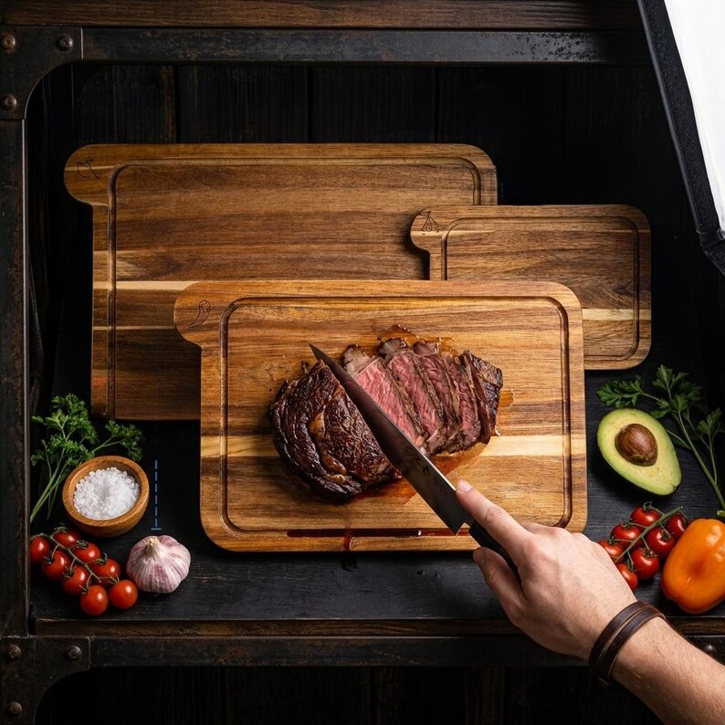 May include: Three wooden cutting boards of varying sizes, with a seared steak being sliced on the front board. The boards are surrounded by ingredients like tomatoes, avocado, and garlic. A hand is holding the knife.