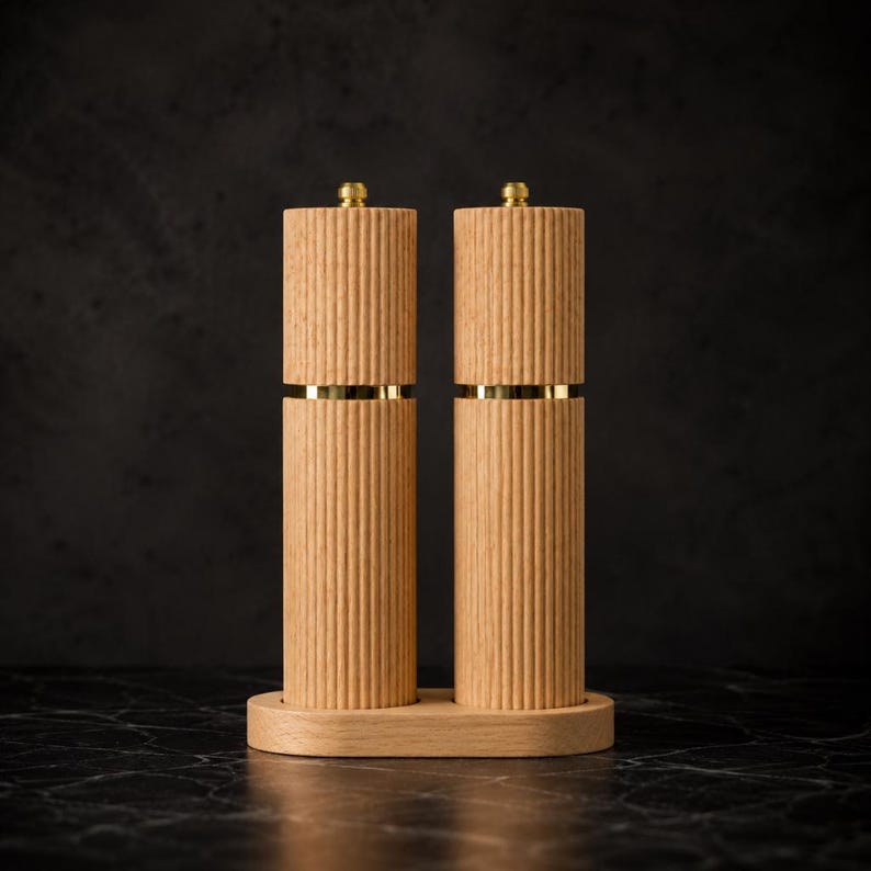 May include: A set of two wooden salt and pepper grinders with a light brown finish and vertical ribbed design. Each grinder has a gold band and a gold top. The grinders sit on a matching wooden base.