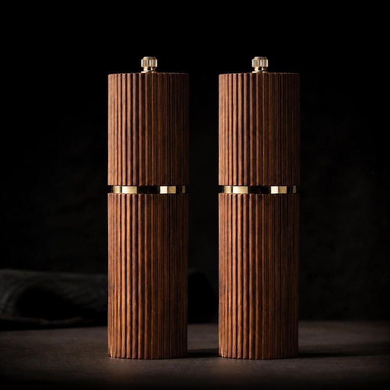 May include: Two wooden salt and pepper grinders with a ribbed design. The grinders are a dark brown color with gold-colored bands and tops. The grinders are cylindrical and stand upright against a dark background.