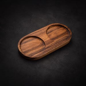 May include: A wooden serving tray with two circular indentations. The tray is oval-shaped and made of a dark brown wood with visible grain patterns. The background is a dark, textured surface.