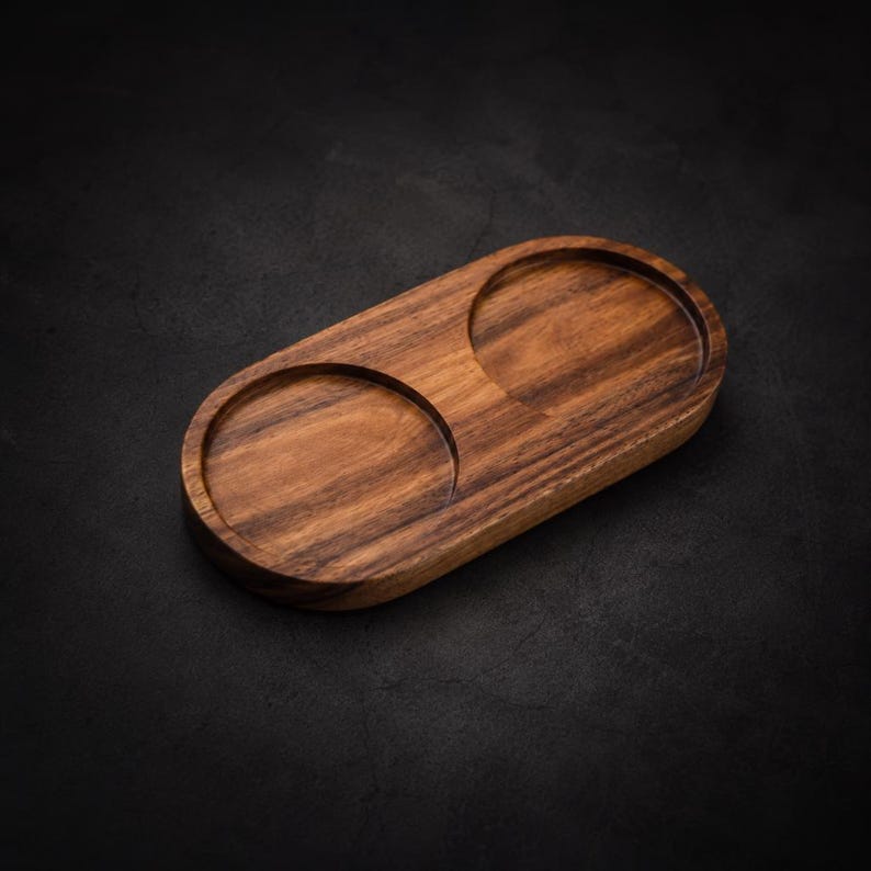 May include: A wooden serving tray with two circular indentations. The tray is oval-shaped and made of a dark brown wood with visible grain patterns. The background is a dark, textured surface.