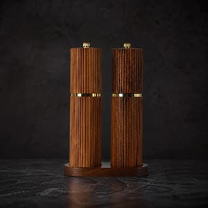 May include: Two wooden salt and pepper grinders with a ribbed design and gold-colored bands. The grinders are displayed on a matching wooden base. The tops of the grinders have a small gold-colored knob.