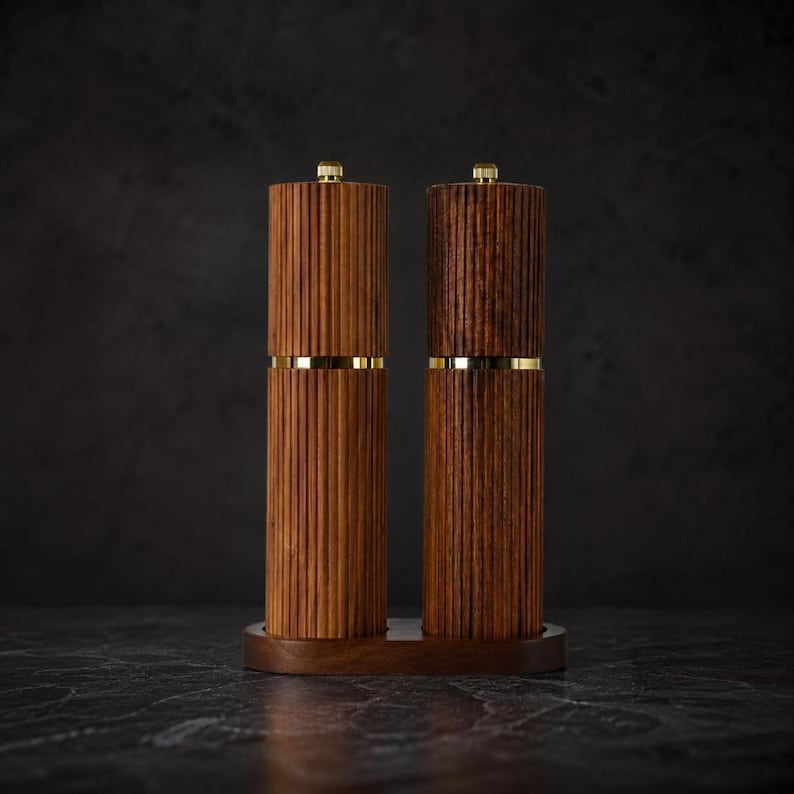 May include: Two wooden salt and pepper grinders with a ribbed design and gold-colored bands. The grinders are displayed on a matching wooden base. The tops of the grinders have a small gold-colored knob.