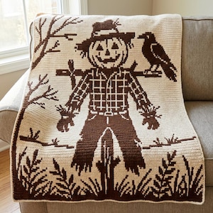 May include: A cream-colored crochet blanket featuring a dark brown graphic design of a scarecrow with a jack-o'-lantern head, wearing a plaid shirt and hat, standing in a field with a crow perched on its shoulder and bare trees in the background.