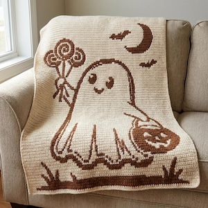 May include: A cream-coloured crochet blanket featuring a brown graphic design of a ghost holding a bouquet of lollipops and a jack-o'-lantern bucket. The scene includes a crescent moon and two small bats above the ghost, with grass at the bottom.