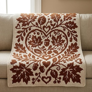 May include: A rectangular crochet blanket featuring a cream-colored base with a repeating pattern of rust-colored autumn leaves, acorns, and heart shapes. The design is centered around a large heart motif composed of intricate botanical elements.
