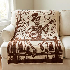 May include: A cream-colored crochet blanket featuring a dark brown graphic design of a skeleton wearing a top hat and playing a banjo while sitting on a wooden fence. The scene includes a bare tree, a crescent moon, stars, and tombstones.