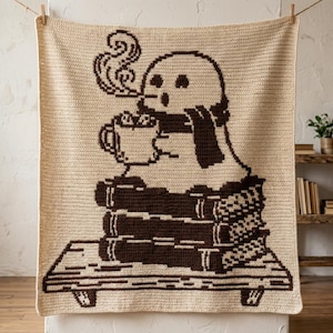 May include: A cream-coloured crochet blanket featuring a dark brown graphic design of a ghost wearing a scarf, holding a steaming mug, and sitting atop a stack of three books on a wooden shelf. The blanket has a rectangular shape and texture.