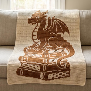 Può includere: Una coperta rettangolare all'uncinetto con un design bicolore crema e marrone. Il centro mostra un grafico dettagliato di un drago appollaiato sopra una pila di tre libri, realizzato con una tecnica di uncinetto arazzo con punti visibili.