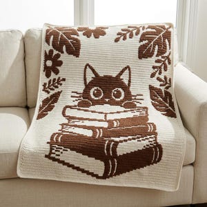 May include: A rectangular crochet blanket featuring a cream-colored base with a dark brown graphic design. The pattern depicts a cat with large eyes peeking over a stack of books, surrounded by a border of various leaves and flower shapes.