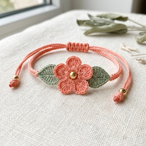 Könnte beinhalten: Handgefertigtes korallenfarbenes Makramee-Armband mit einer gehäkelten Blume mit goldenem Zentrum und zwei grünen Blättern. Das Armband hat verstellbare Kordeln und goldfarbene Perlen an den Enden. Ein zartes Blumenaccessoire.
