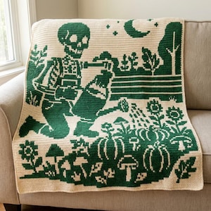 May include: A cream-colored crochet blanket featuring a dark green graphic design of a skeleton kneeling and watering a garden. The scene includes sunflowers, pumpkins, mushrooms, a crescent moon, and stars, all rendered in a detailed knit pattern.