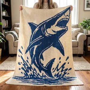 May include: A rectangular crochet blanket featuring a large, dark blue graphic of a shark leaping out of water against a cream-colored background. The design shows the shark with its mouth open, surrounded by stylized blue splashes at the bottom.